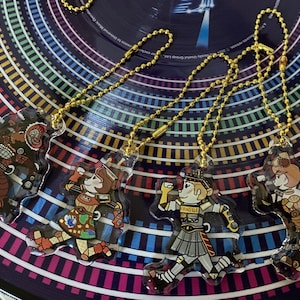 May include: Four colorful cartoon character keychains with gold chains. The characters are wearing different outfits and have different expressions. The keychains are on a colorful background.