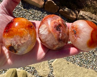 Puede incluir: Tres piedras de cornalina pulidas con una mezcla de tonos naranja, rojo y blanco. Las piedras en forma de huevo se sostienen en una mano, mostrando sus patrones y texturas naturales. Estas piedras son ideales para coleccionistas de cristales.