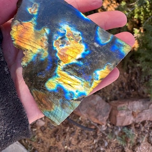 Può includere: Un grande cristallo di labradorite iridescente con un bagliore blu, verde e dorato. Il cristallo è grezzo e ha una forma naturale irregolare.