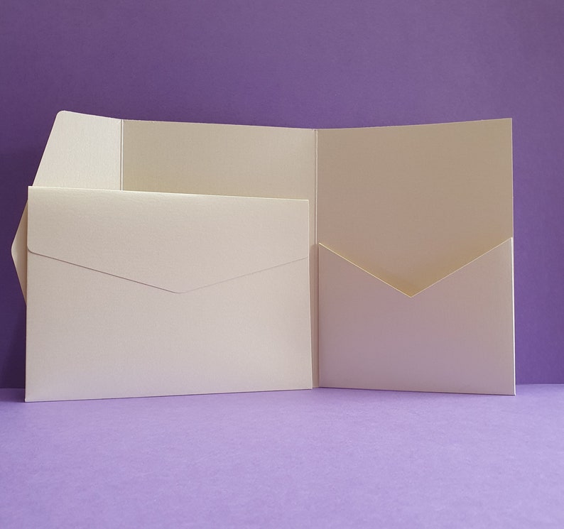 K&ouml;nnte beinhalten: Ein wei&szlig;er Umschlag mit einer Klappe auf der Vorderseite und einer Tasche auf der R&uuml;ckseite. Der Umschlag ist in der Mitte gefaltet und liegt auf einer violetten Oberfl&auml;che.