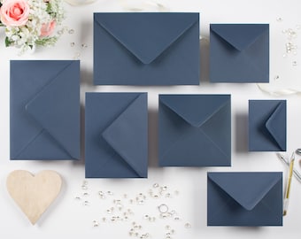 May include: A collection of navy blue envelopes in various sizes, arranged on a white surface. A wooden heart, a bouquet of flowers, and scattered decorative elements complement the stationery. Ideal for wedding invitations or announcements.