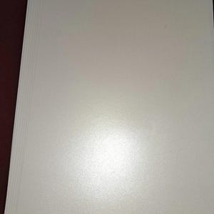 May include: A pile of white, textured paper sheets. The paper has a subtle sheen and a slightly rough surface. The edges of the paper are visible, showing the thickness of the stack. The paper appears to be clean and unused.