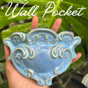 May include: A blue ceramic wall pocket with raised, cream-colored scrollwork details. The text "slip-Casted Wall Pocket" is printed at the top, and "@LateBloomer.PlantCo" is printed at the bottom of the image.
