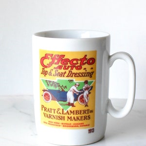 Może przedstawiać: Biały kubek ceramiczny z vintage'ową reklamą "Effecto Auto Top & Seat Dressing" firmy Pratt & Lambert Varnish Makers. Reklama przedstawia samochód i dwie osoby w strojach z epoki.