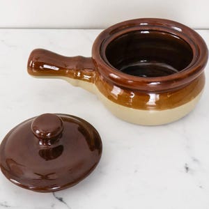 May include: A two-tone ceramic soup bowl with a handle and a lid. The bowl is brown and beige, with a dark brown interior and lid. The lid is off to the side, revealing the inside of the bowl.