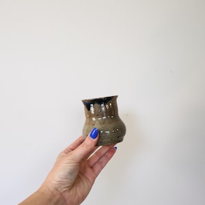 May include: A small, hand-thrown ceramic cup with a bulbous body and a flared rim. The vessel features a speckled, earthy brown glaze with a dark, drippy black glaze accenting the upper rim, showcasing a smooth, glossy finish throughout.