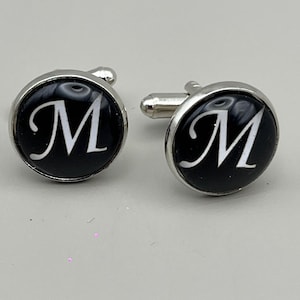 May include: A pair of silver cufflinks with a black background and the white letter "M" in a cursive font. The cufflinks are round with a shiny silver border and a post on the back.