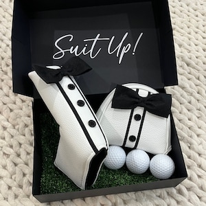 Puede incluir: Una caja de regalo negra con las palabras "Suit Up!" impresas en la tapa. La caja contiene dos cubrecabezas de golf blancos con pajaritas negras y cuatro bolas de golf blancas.