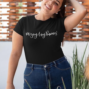 May include: Black t-shirt with the text "raising tiny humans" in a cursive font. The model is wearing dark blue jeans. The background is a brick wall.