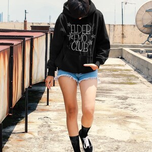May include: Black hoodie with the text "ELDER EMO CLUB" in white, paired with denim shorts and black sneakers. The hoodie features a drawstring hood and a kangaroo pocket. The outfit is completed with black socks.
