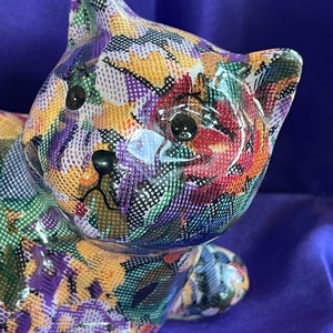 Op de afbeelding: Een keramiek kattenbeeldje met een kleurrijk bloemenpatroon. De kat ligt met zijn kop naar de zijkant gedraaid. Het beeldje is geschilderd in tinten paars, groen, geel en rood.