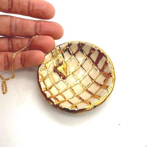 May include: A small, round, cream-colored dish with a gold grid pattern and gold trim. A gold necklace with a star pendant rests inside the dish. The dish is held by a hand against a white background.