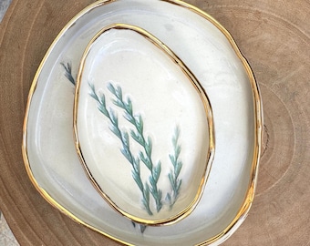 Puede incluir: Dos platos de cerámica, uno dentro del otro. Los platos son de color blanquecino con un borde dorado. El plato más pequeño tiene un borde dorado y una ramita pintada de hojas verde azulado. El plato más grande tiene un diseño similar.