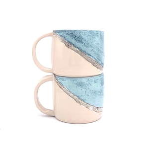 May include: Two stacked ceramic mugs with a light blue and white glaze. The mugs have a diagonal stripe design with a light brown accent.