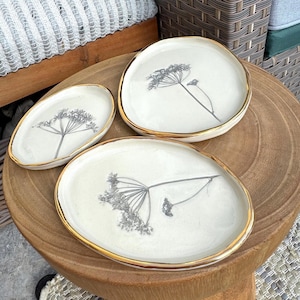 Puede incluir: Tres platos de cerámica color crema con borde dorado. Cada plato presenta un diseño botánico gris. Los platos están dispuestos sobre una superficie de madera, mostrando un conjunto de fuentes decorativas.