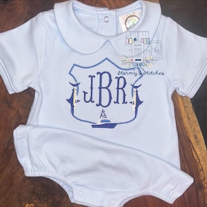 Puede incluir: Romper de bebé azul claro con cuello Peter Pan y mangas cortas. El romper presenta un escudo bordado en azul marino con las iniciales "JBR" y un diseño de barco. También es visible el texto "Stormy's Stitches".