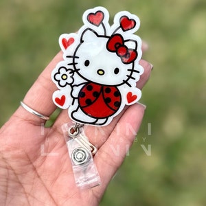 May include: A white Hello Kitty character with red ladybug wings and a red bow. She is holding a white flower and has red hearts around her. The character is on a clear plastic badge holder with a silver clip.