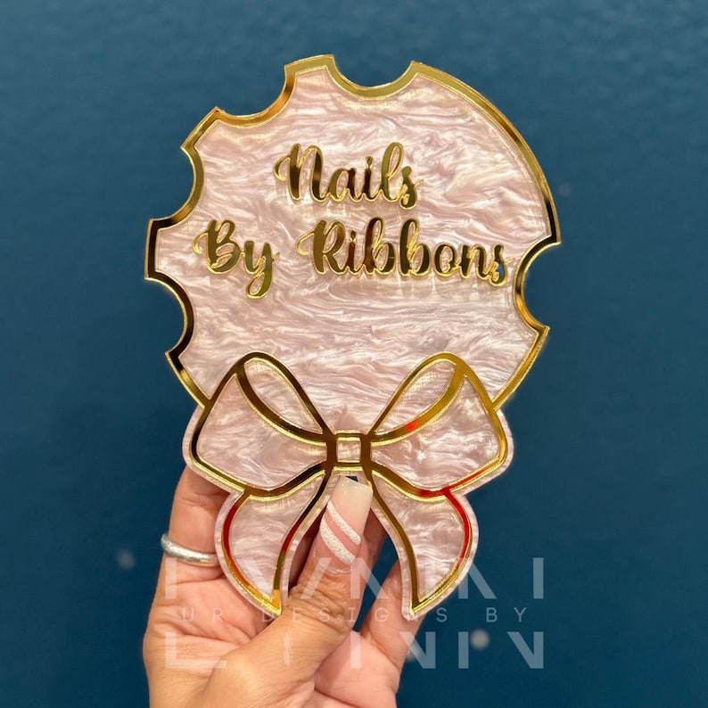 Pode incluir: Uma placa de acr&iacute;lico rosa e dourado com um la&ccedil;o e o texto "Nails By Ribbons".