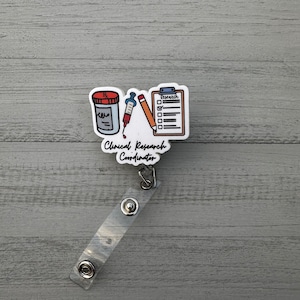May include: White badge holder with a cartoon illustration of a pill bottle, syringe, pencil, and clipboard with the text "Research" on it. The text "Clinical Research Coordinator" is below the illustration. The badge holder has a clear plastic strap with a metal clip.