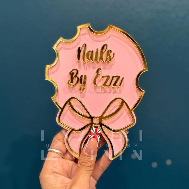 Pode incluir: Um letreiro de acr&iacute;lico rosa e dourado com um la&ccedil;o e o texto "Nails By Ezz" em letras douradas.