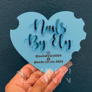 May include: A light blue heart-shaped acrylic sign with the text "Nails By Ely" in a white, reflective font. The sign also includes social media handles: @nailsbyely2024 and @nails.by.ely.2024.