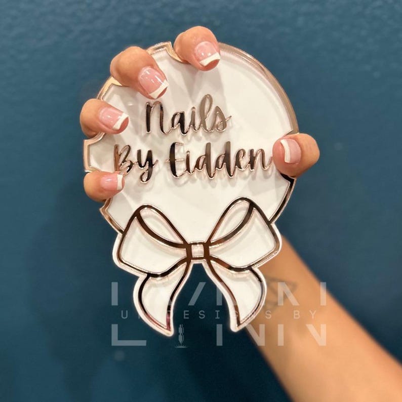 Pode incluir: Um sinal redondo branco com um la&ccedil;o dourado e o texto "Nails by Fidden" em letras douradas. Uma m&atilde;o com unhas brancas com manicure francesa est&aacute; segurando o sinal.