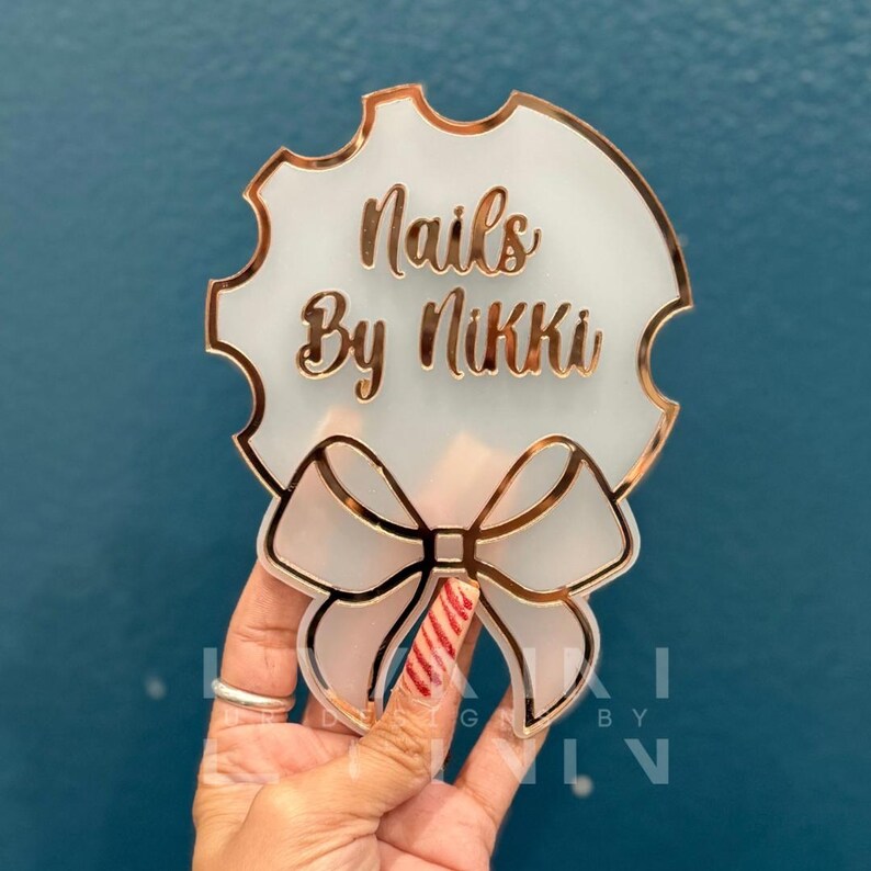 Pode incluir: Um letreiro de acr&iacute;lico branco com contorno dourado e um la&ccedil;o. O letreiro diz "Nails By Nikki".