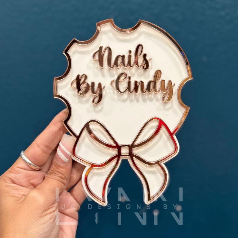 Pode incluir: Um letreiro de acr&iacute;lico branco com uma borda e um la&ccedil;o de ouro rosa. O letreiro diz "Nails By Cindy".