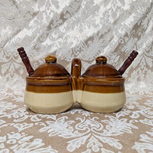 May include: A brown and white ceramic double honey pot with wooden dippers. The honey pots have a brown glaze with a white band around the middle.