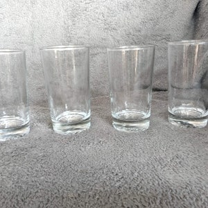 May include: Four clear glass tumblers on a gray textured surface. The tumblers are all the same size and shape.