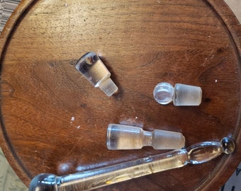 Puede incluir: Cuatro tapones de botella de vidrio transparente sobre una superficie de madera. Los tapones son de varias formas y tamaños. Un tapón tiene un bulbo redondo en el extremo de un tallo largo y delgado.