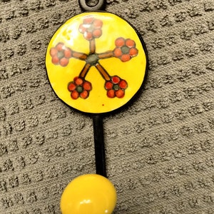 May include: A decorative wall hook. It has a yellow circular design with red and grey flower-like shapes. The hook has a black stem and a yellow knob at the bottom. The top has a black ring for hanging.
