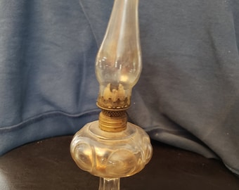 May include: A clear glass antique oil lamp with a decorative base and a broken chimney. The lamp has a brass burner and a round, patterned reservoir. The lamp is likely from the early 20th century.
