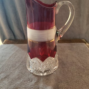 May include: A ruby red glass jug with a clear handle. The jug features a white band with gold trim. The base of the jug has a cut glass design. The jug is likely used for serving beverages.
