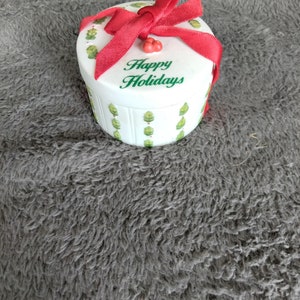 May include: A white ceramic gift box with a red ribbon and green holly leaves. The box has the words "Happy Holidays" written on it.