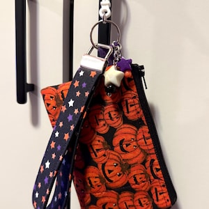 May include: A fabric wristlet pouch featuring an orange pumpkin pattern on a black background with a matching wrist strap decorated with small stars. The pouch includes a black zipper, a silver metal clasp, and a white plastic clip with charms.