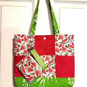 May include: A patchwork tote bag featuring a pattern of watermelon slices, strawberries, and cherries on a white background, accented with solid red polka-dot panels and a green batik-style base. A matching small zippered pouch is attached.