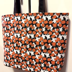 May include: A rectangular fabric tote bag featuring a repeating pattern of orange pumpkins and white ghosts on a black background. The bag has two black shoulder straps with visible orange stitching, providing a sturdy handle for carrying items.