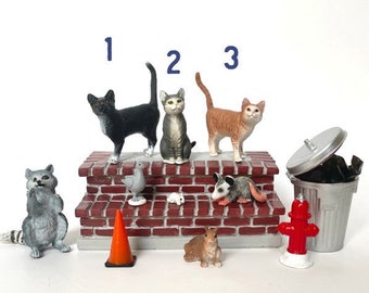 Könnte beinhalten: Eine Sammlung von Miniaturfiguren zum Thema Stadt, darunter drei Katzen, ein Waschbär, ein Eichhörnchen, ein Opossum, eine Taube, eine Maus, ein Verkehrskegel, ein Hydrant und ein Mülleimer. Der Text lautet "THE ORIGINAL CITY PACK + 1 CAT OF YOUR CHOICE."