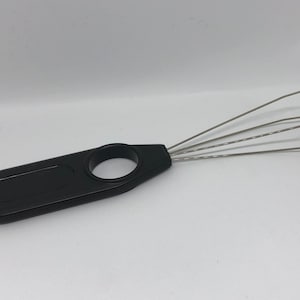 Könnte beinhalten: Ein schwarzes Plastikwerkzeug mit einem Ringgriff und fünf dünnen Metalldrähten, die sich vom Griff erstrecken. Die Drähte sind zu einer Form gebogen, die einem Rechen ähnelt.