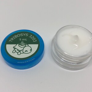 May include: A small, clear jar filled with white cream, next to a blue lid. The lid has the text "TRIBOSYS 3203" and a cartoon turtle graphic. The jar contains 5 mL of product.