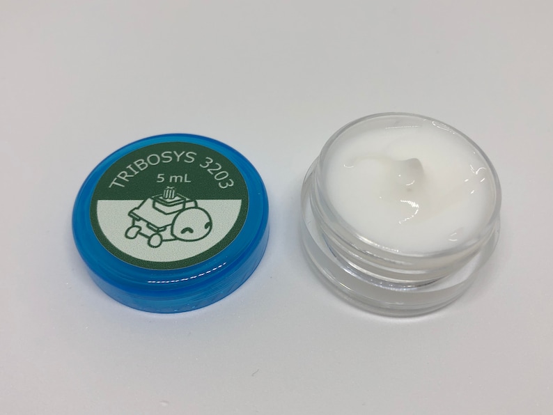 May include: A small, clear jar filled with white cream, next to a blue lid. The lid has the text "TRIBOSYS 3203" and a cartoon turtle graphic. The jar contains 5 mL of product.