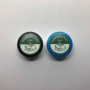 May include: Two small, round containers with lids. One lid is black, the other is blue. Both lids have the text "TRIBOSYS 3203" and a graphic of a turtle on a machine. The black container is labeled "3 mL", the blue one "5 mL".