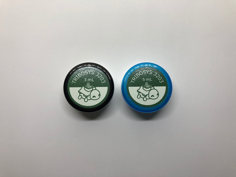 May include: Two small, round containers with lids. One lid is black, the other is blue. Both lids have the text "TRIBOSYS 3203" and a graphic of a turtle on a machine. The black container is labeled "3 mL", the blue one "5 mL".