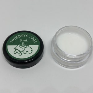 May include: A small, clear plastic jar filled with white lubricant, next to a black lid. The lid has a green label with the text "TRIBOSYS 3203" and a cartoon image of a robot. The jar contains 3 mL of lubricant.