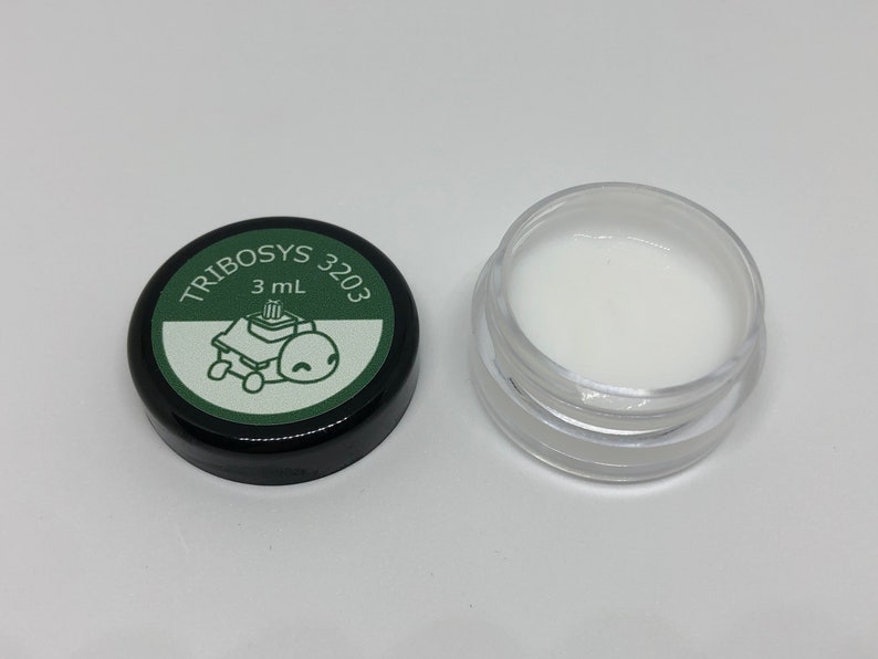 May include: A small, clear plastic jar filled with white lubricant, next to a black lid. The lid has a green label with the text "TRIBOSYS 3203" and a cartoon image of a robot. The jar contains 3 mL of lubricant.