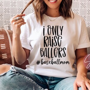 May include: White t-shirt with black text that reads "I only raise ballers #baseballmom".