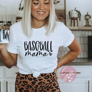 May include: White t-shirt with black text that reads "Baseball mama" with a small heart symbol. The person wearing the shirt is smiling and holding a black and white mug.
