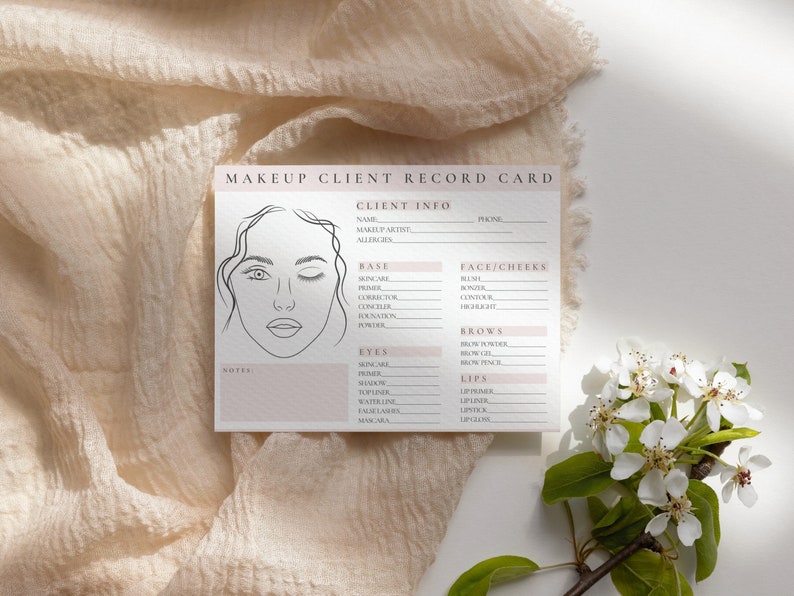 May include: A makeup client record card with a line drawing of a woman's face. The card has sections for client information, base makeup, eyes, face and cheeks, brows, and lips. The card is on a white surface with a white flower.