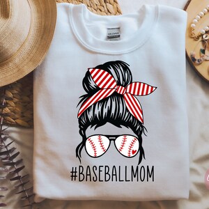 May include: White sweatshirt featuring a graphic of a woman with a messy bun wearing baseball-themed sunglasses and a red and white striped headband. The text "#BASEBALLMOM" is printed below the graphic.